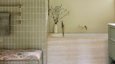 green bathroom with travertine built-in bath with vase of flowers, and gold taps. there's gold towel rails on a wall of green tiles, and a bench seat below with a patterned seat
