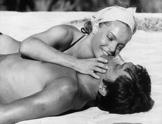 Romy Schneider laying by the pool in a black bikini and white bandana while filming La Piscine (The Swimming Pool) with Alain Delon.