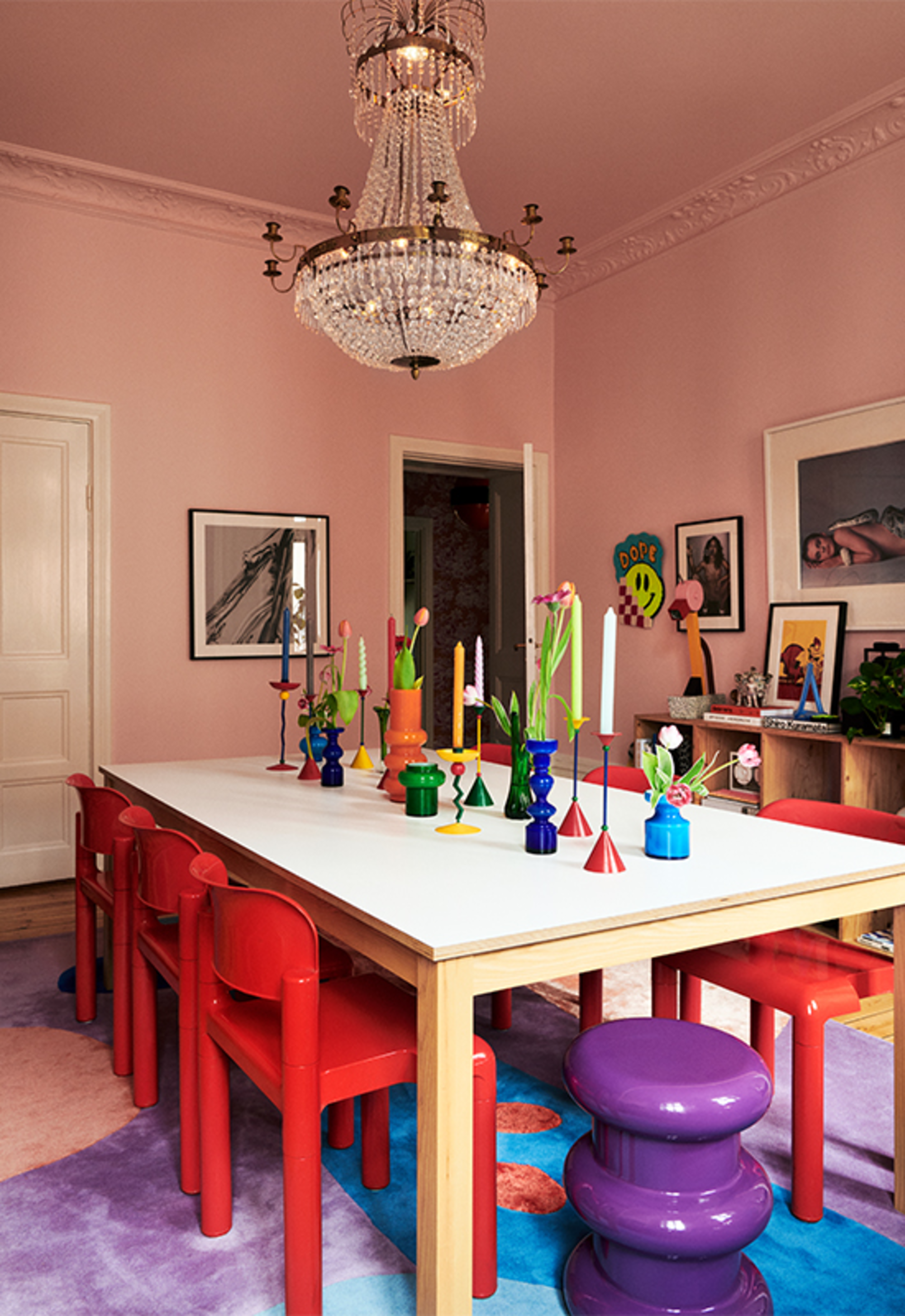 colorful dining room with scandi style with colorful rug, purple stool, red dining chair, pink walls and ceiling, chandelier, and colorful candlesticks on the dining table