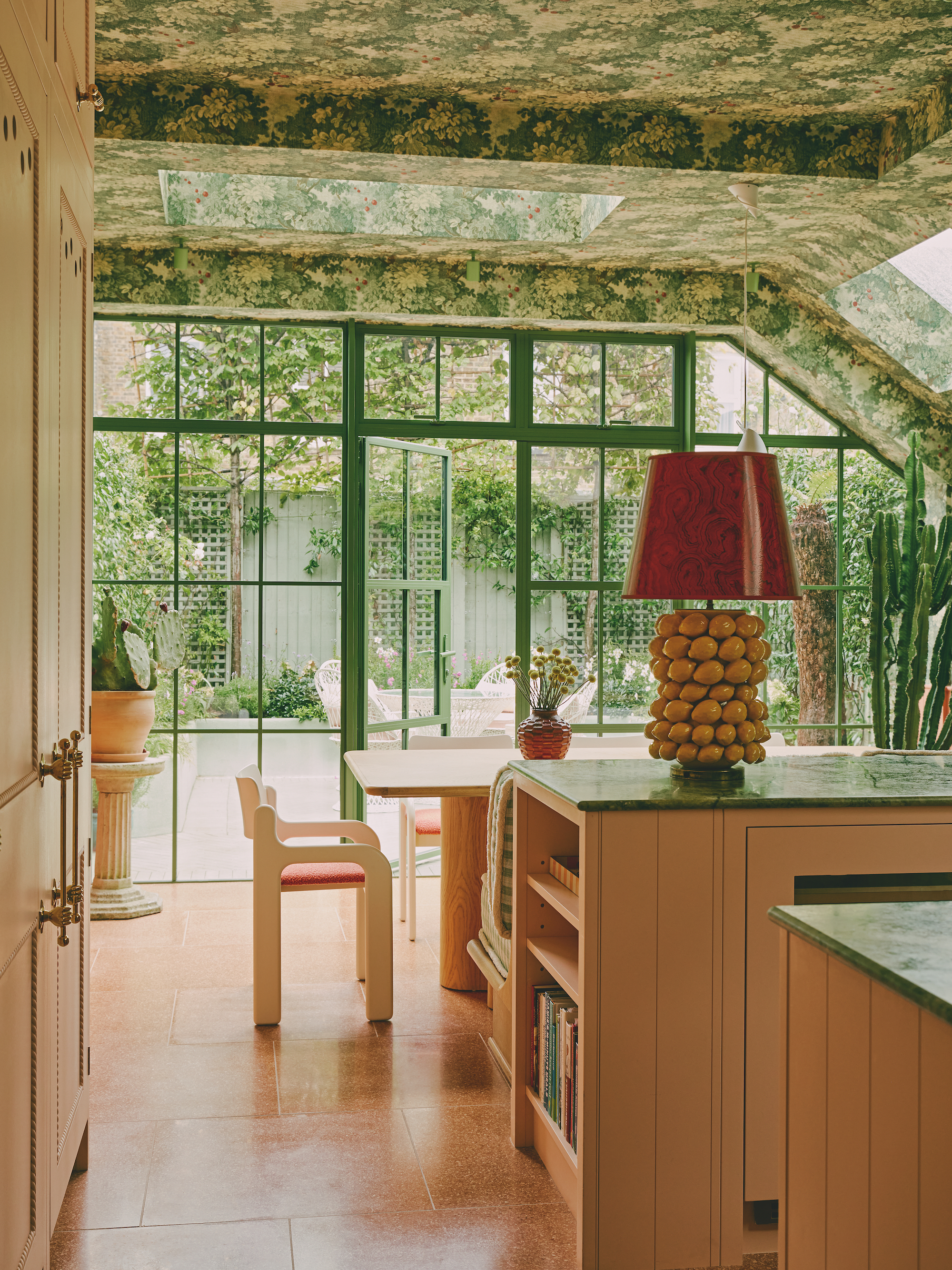 A modern, green filled kitchen with wallpapered ceilings, green worktops and a modern cream dining table. The glass doors to the outdoor space are lined with green painted metal and the floor tiles are a peachy orange shade.