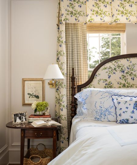 Bedroom with cream walls, a window with a bamboo roman blind, a floral bed canopy with gingham drapes, a mahogany bed upholstered in a floral print, and a nightstand with books and a sconce above it.
