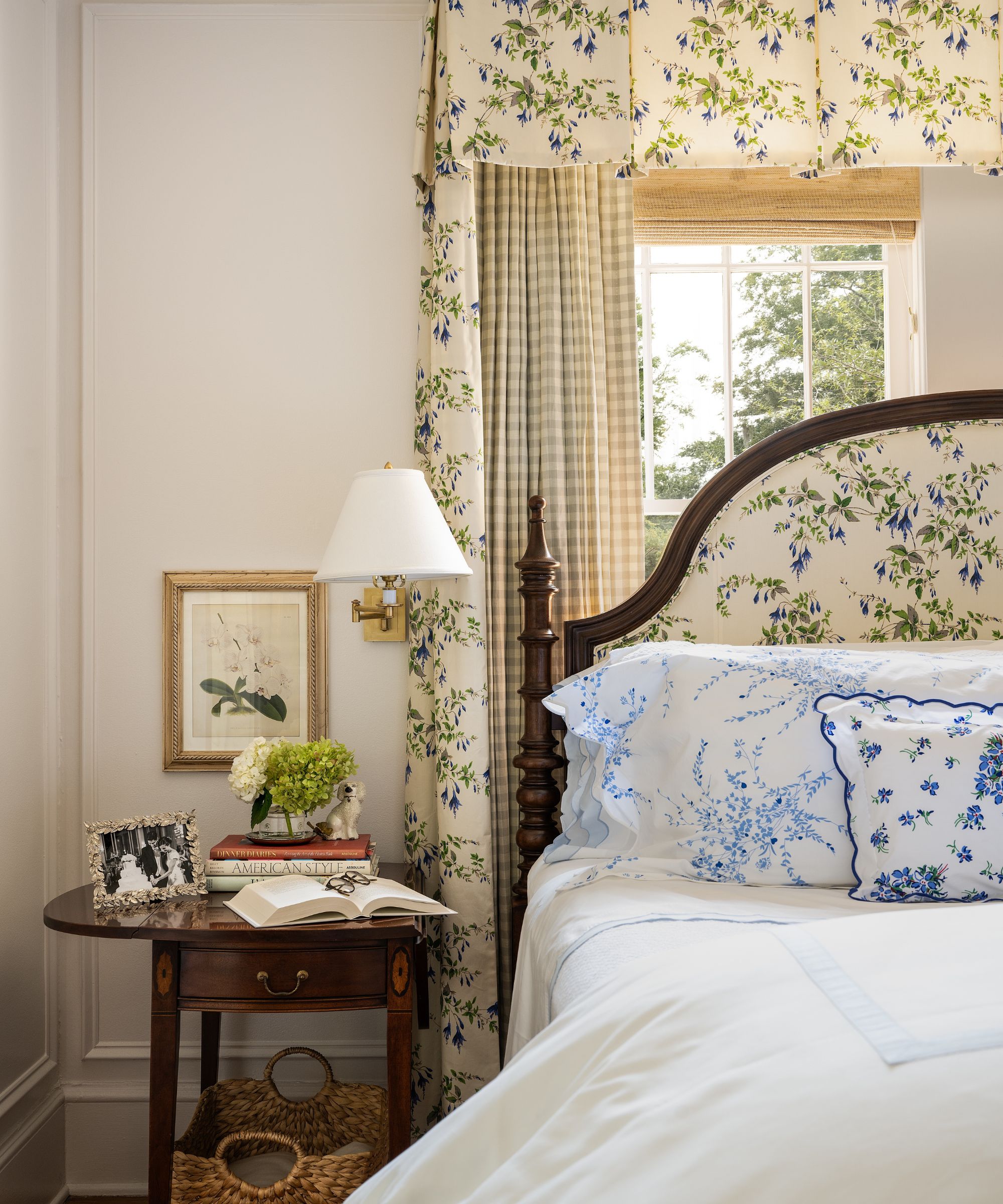 Bedroom with cream walls, a window with a bamboo roman blind, a floral bed canopy with gingham drapes, a mahogany bed upholstered in a floral print, and a nightstand with books and a sconce above it.