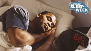 A man lies asleep in bed next to a black digital alarm clock displaying a time of 6.23am