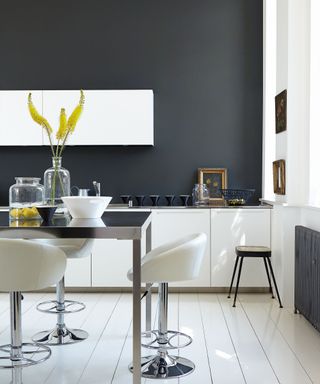 Kitchen space with graphite painted wall, white flooring and kitchen cupboards, high kitchen island in metal and black, white and metal high bar stools, decorative vases and ornaments on table, low stool beside gray radiator, decorative picture frames and paintings on wall and resting on countertop