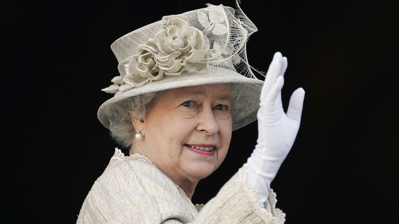 Queen Elizabeth wearing a beige hat with flowers and matching coat with white gloves and waving against a black background 