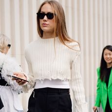 A street style photo from London Fashion Week showing a guest wearing an elegant cream cropped sweater with black Bermuda shorts and strappy heeled sandals.