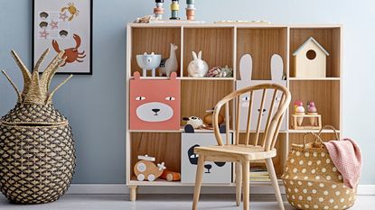 A blue children's bedroom with a wooden square cubby bookcase filled with toys. A wooden chair in front of it, and a basket for blankets 