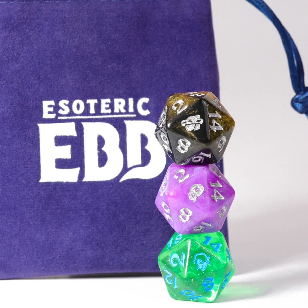 Esoteric Ebb three standard dice stacked up next to purple bag with game name