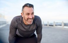 An attractive, fit older man leans on his knees and looks happily off-camera. He may be looking a his dog or taking a break from running.