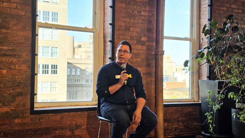 A man holding a microphone sits on a stool in a warmly lit room with exposed brick walls and large windows. He appears engaged and thoughtful.