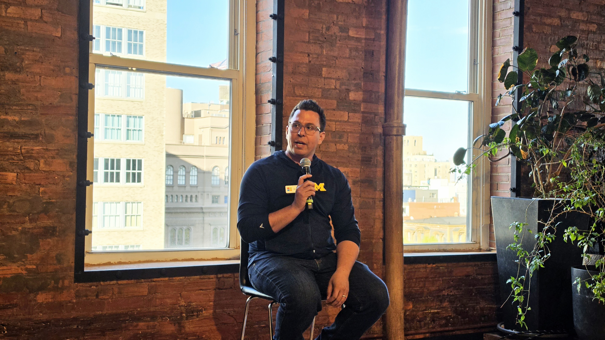 A man holding a microphone sits on a stool in a warmly lit room with exposed brick walls and large windows. He appears engaged and thoughtful.