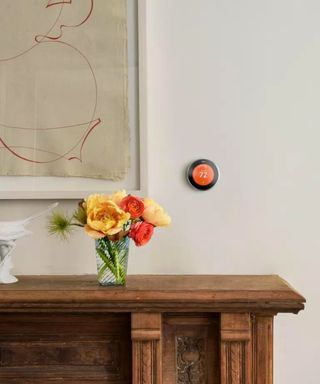 A small orange smart thermostat on a beige wall above a brown sood side boared with red and yellow flowers