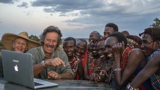 Jonathan Scott in a khaki shirt sits at a table outside with his wife Angela in a straw hat with members of the Maasai wearing traditional jewellery. They smile at something they are watching on a laptop.