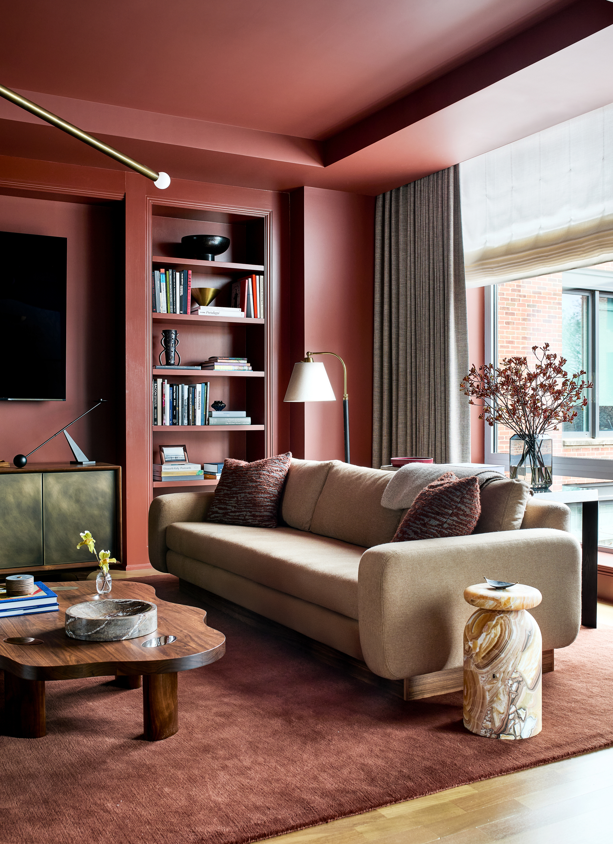 living room color-drenched in red with built-in bookcase, timber wavy coffee table, red rug, stone side table, and beige sofa with patterned curtains