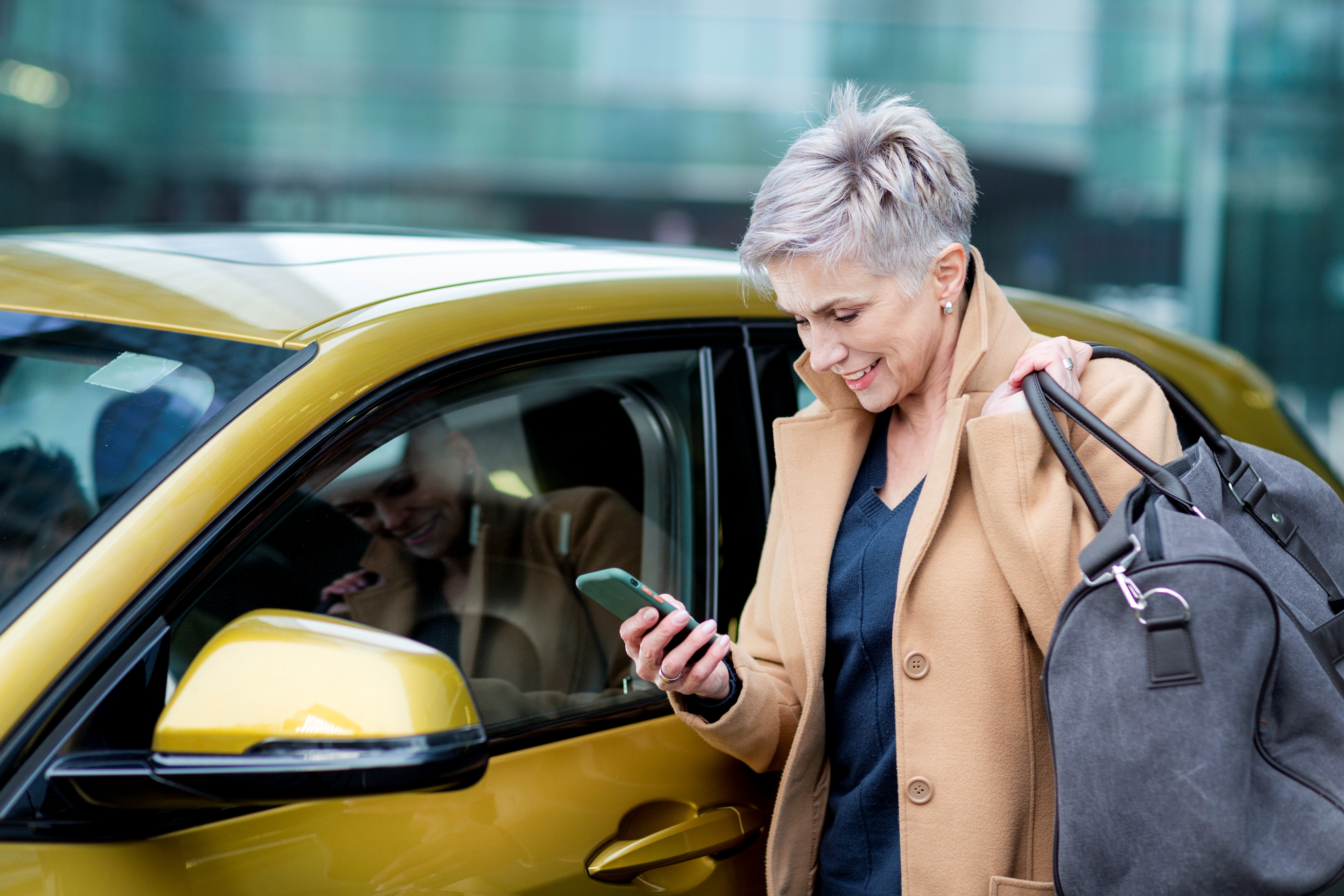 A senior woman with a duffel bag checks her phone before getting into a car.