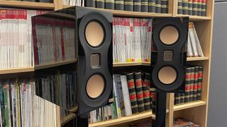 A hands-on photo of the black and gold Studio 89 speakers in front of bookcases
