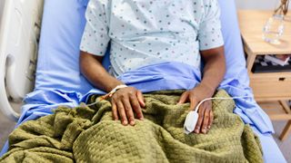 Cropped image of a patient sat in a blue hospital bed where only their mid-section can be seen. They are wearing a polka-dot gown and are covered with a green blanket.