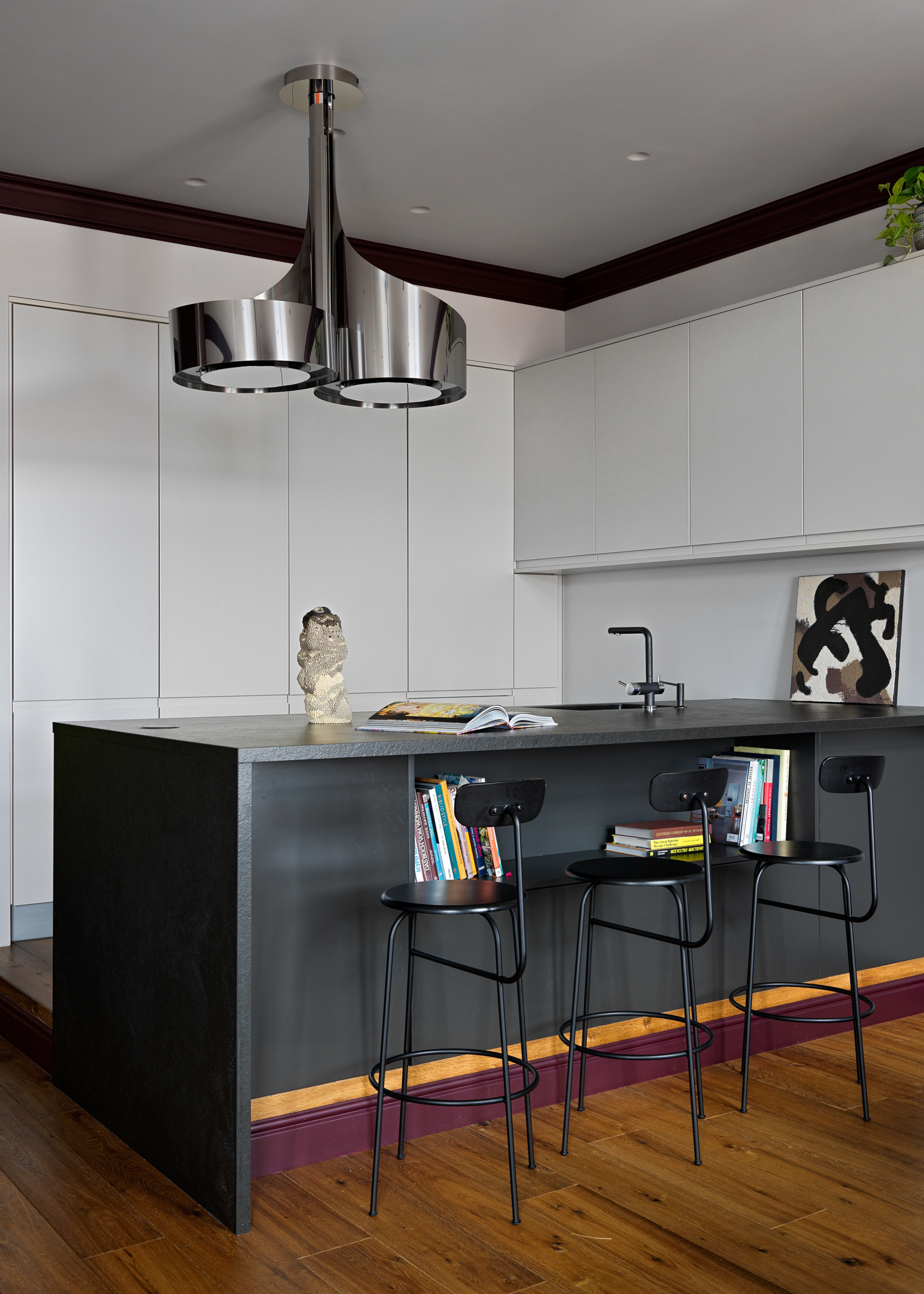 A kitchen with a dark island, black stools, a leaning painting, warm gray cabinetry, and a chrome pendant light