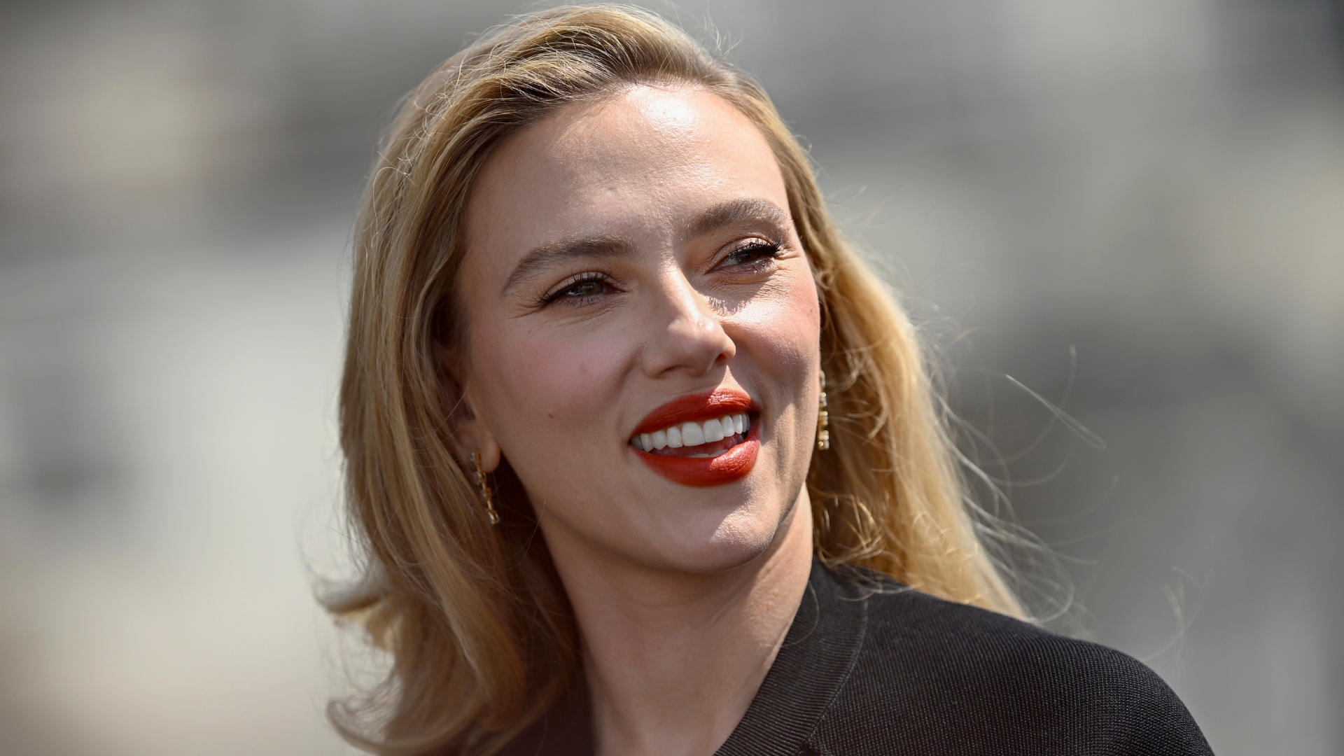 Scarlett Johansson with side-swept blonde hair and wearing a coral red lipstick, smiling as she looks over her shoulder