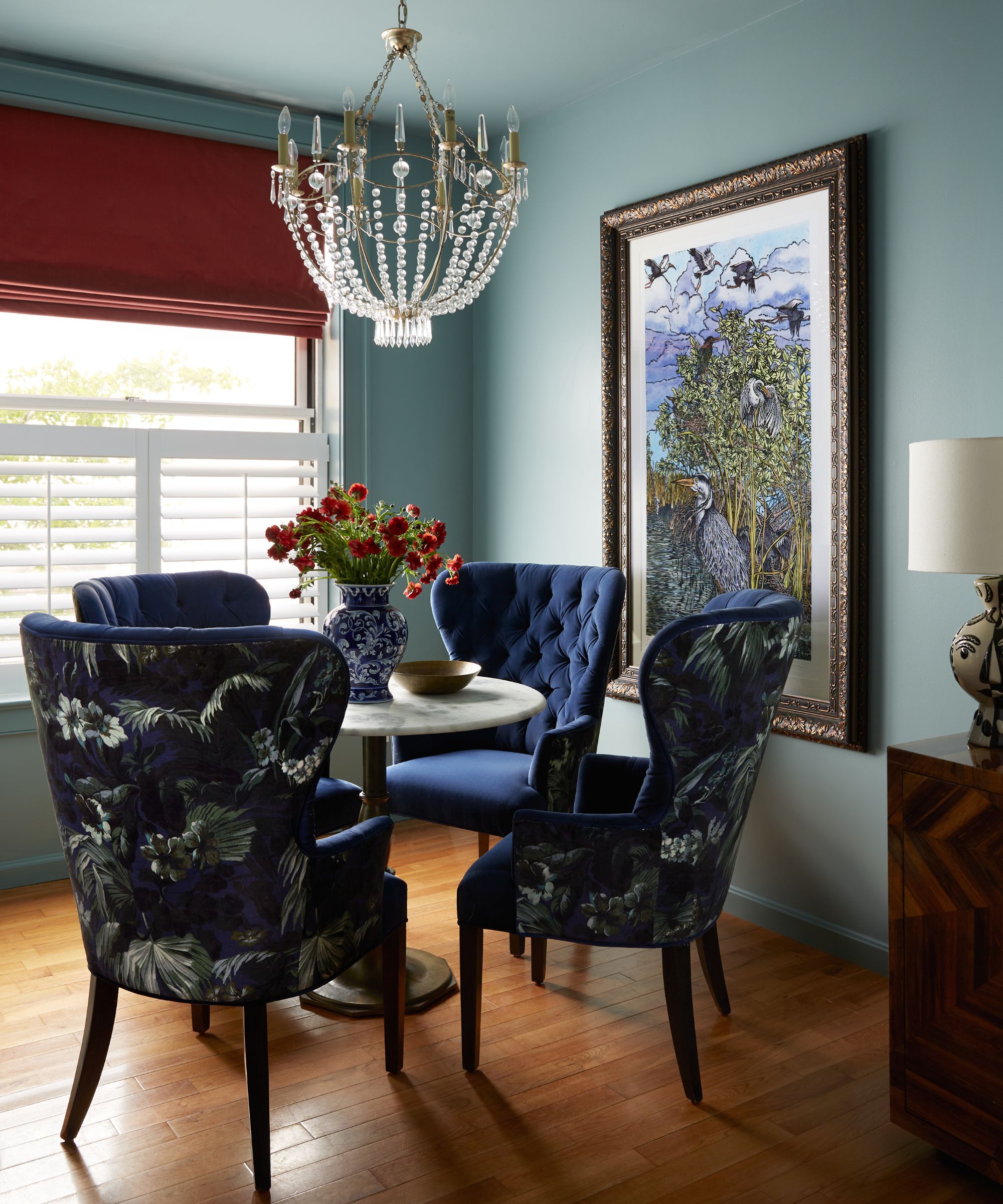 A dining nook with blue-gray walls and ceiling, wood flooring, a round dining table with four dark blue upholstered chairs, and a window with shutters and a red roman blind.