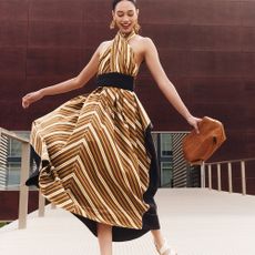 Model wearing a patterned dress sold at The Outnet holding a brown clutch bag