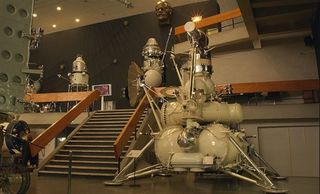 A model of the Lunar 24 moon sample return spacecraft in the center of the image next to a staircase.