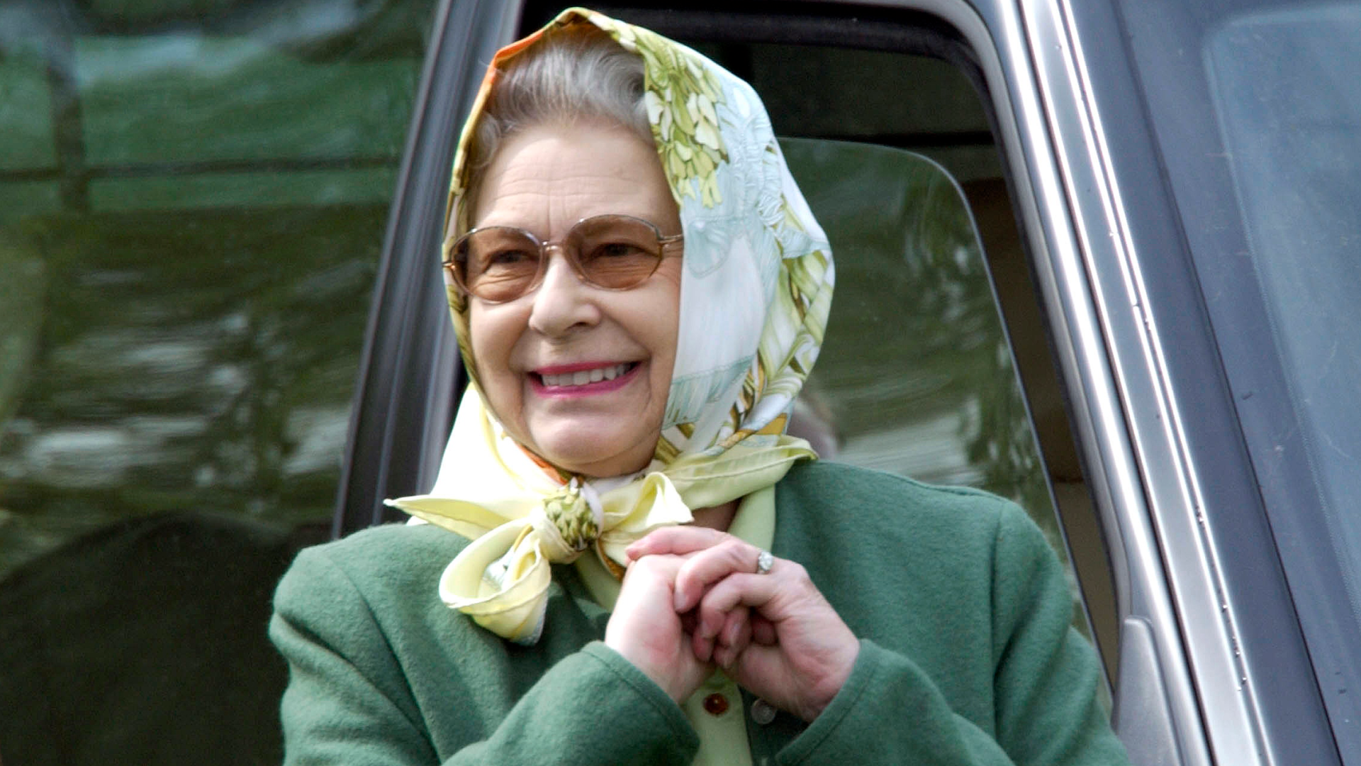 Queen Elizabeth wearing a head scarf and a green coat smiling in front of a car