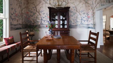 a pink floral mural wallpapered dining room with half height paneling, dark wooden antique furniture and a window bench
