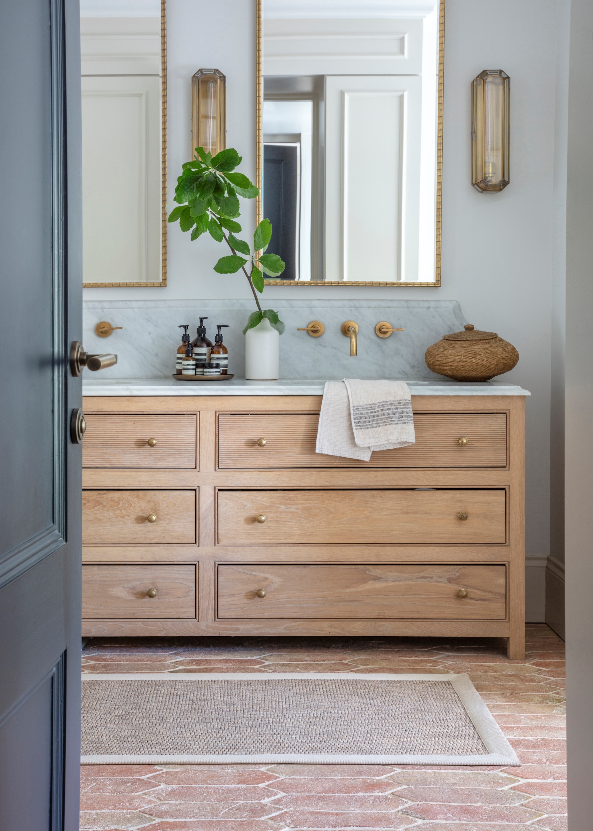 A guest bathroom with a vanity that has multiple drawers. On top of the vanity is a tray of body wash/ soap as well as a plant in a vase and a small rattan basket. Above the vanity are two large bobbin mirrors.