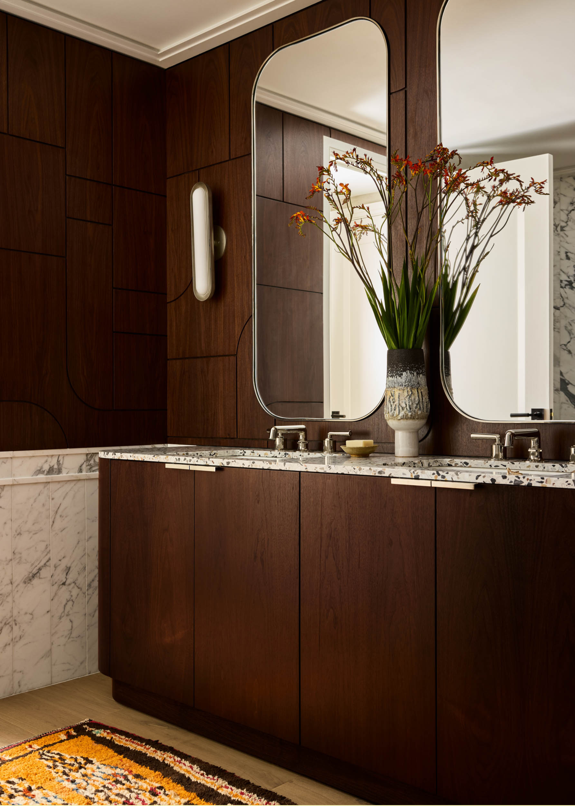 A bathroom with his and hers terrazzo sinks, wood cabinets, a vase of flowers, and a patterned bath mat
