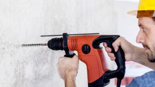 Man in yellow safety helmet using an orange SDS drill to drill into concrete wall