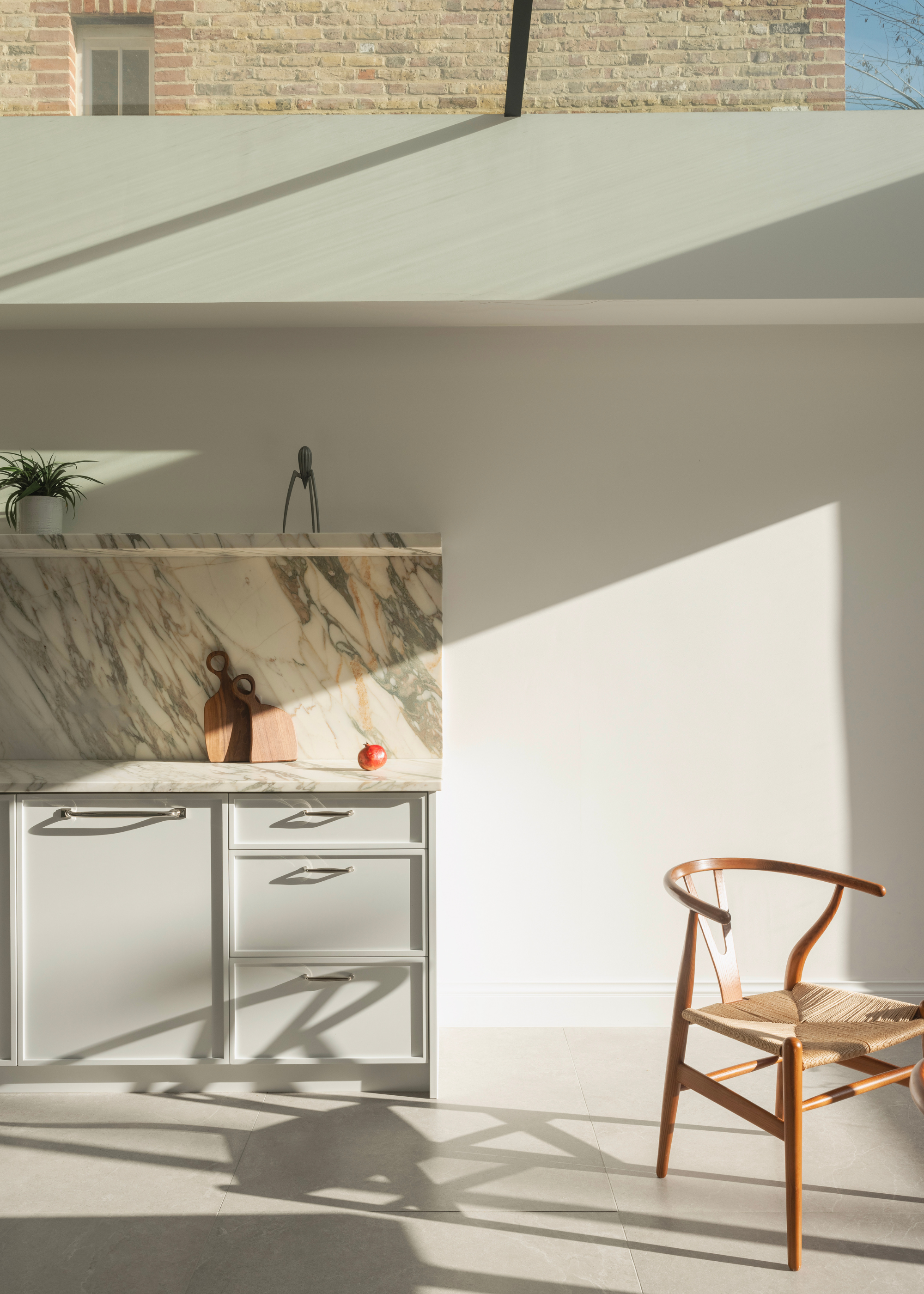 A kitchen with light pouring in and onto a marble kitchen backsplash with a potted plant, a pair of wooden cutting boards and a fruit