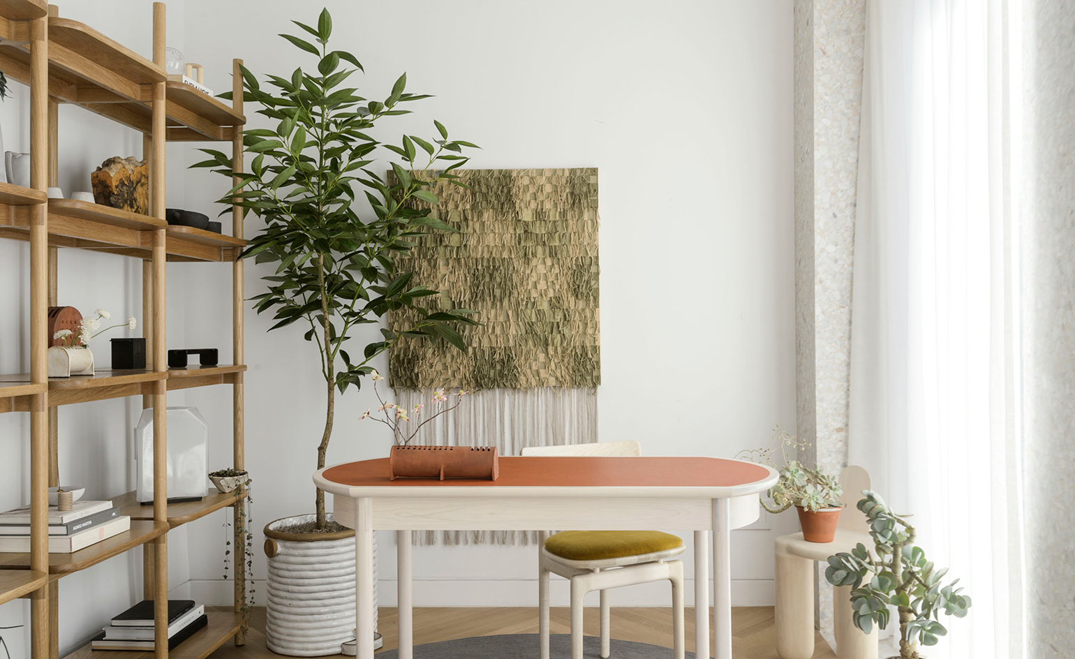 A wood-style bookshelf on the left, next to a white table in the middle with a brown top, a large indoor plant and a white chain.