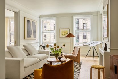 a white apartment living room with neutral furniture looking out of two large windows, a pocket door leads to another room behind a white sofa