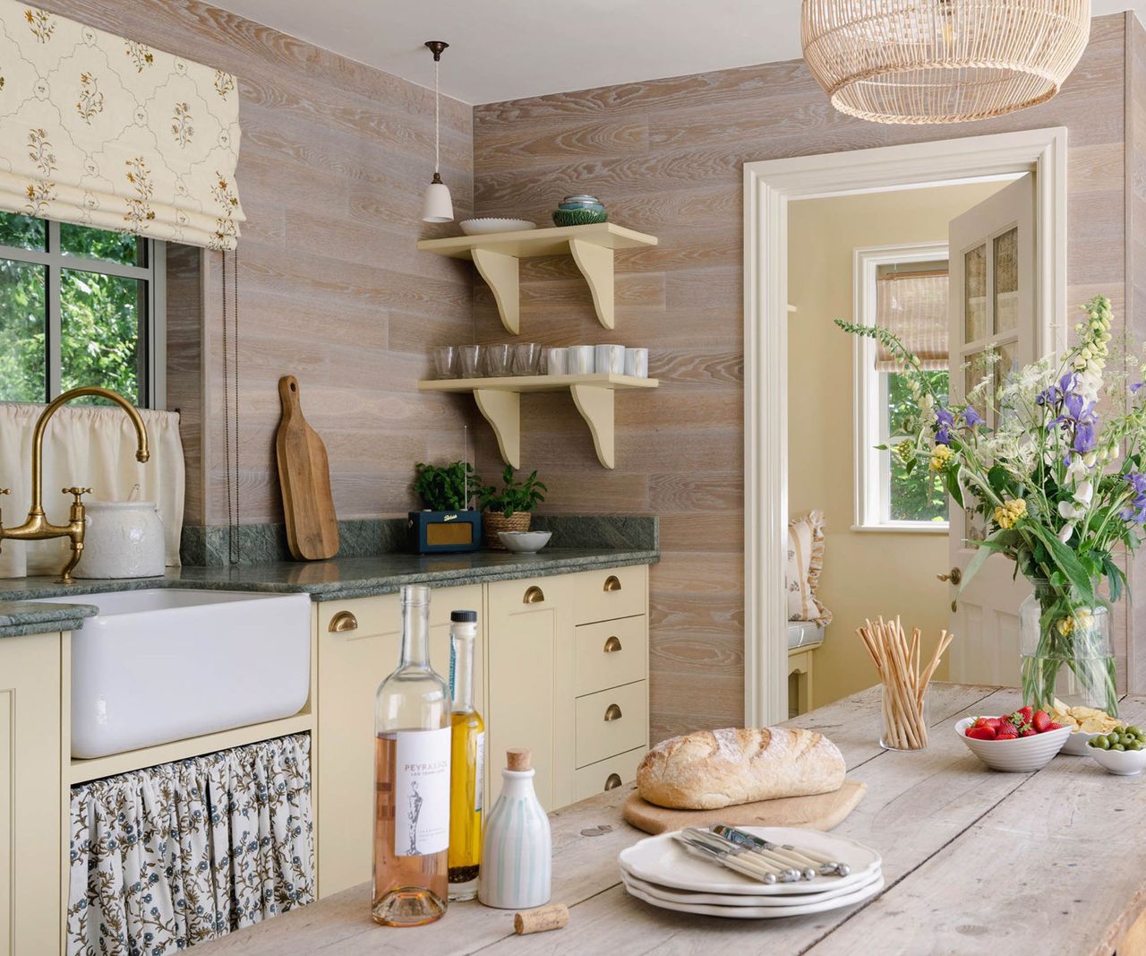 Cream country kitchen, wood-panelled wall, green worktop, wildflowers, bread and wine on rustic table.