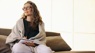 Woman writing in journal, sitting on sofa with her legs up looking into the distance