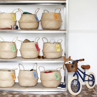 9 soft baskets with colourful labels, 3 per shelf on a large silver shelving unit with wooden shelves, with a blue kids bike with a brown basket leaning up next to it