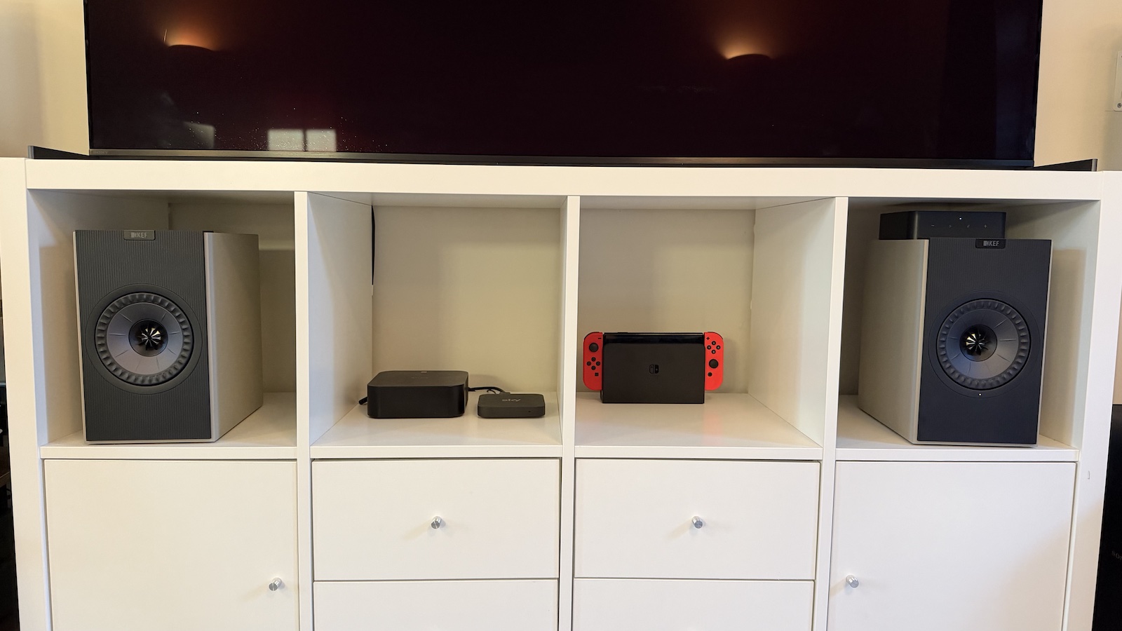 A white bookcase, upon which is standing an OLED TV, and in the nooks of which are a pair of KEF Coda W speakers, a Nintendo Switch, and a couple of other smaller components
