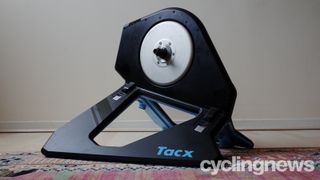 A black Tacx Neo 2T smart trainer stands on a coloured carpet in front of a blank wall