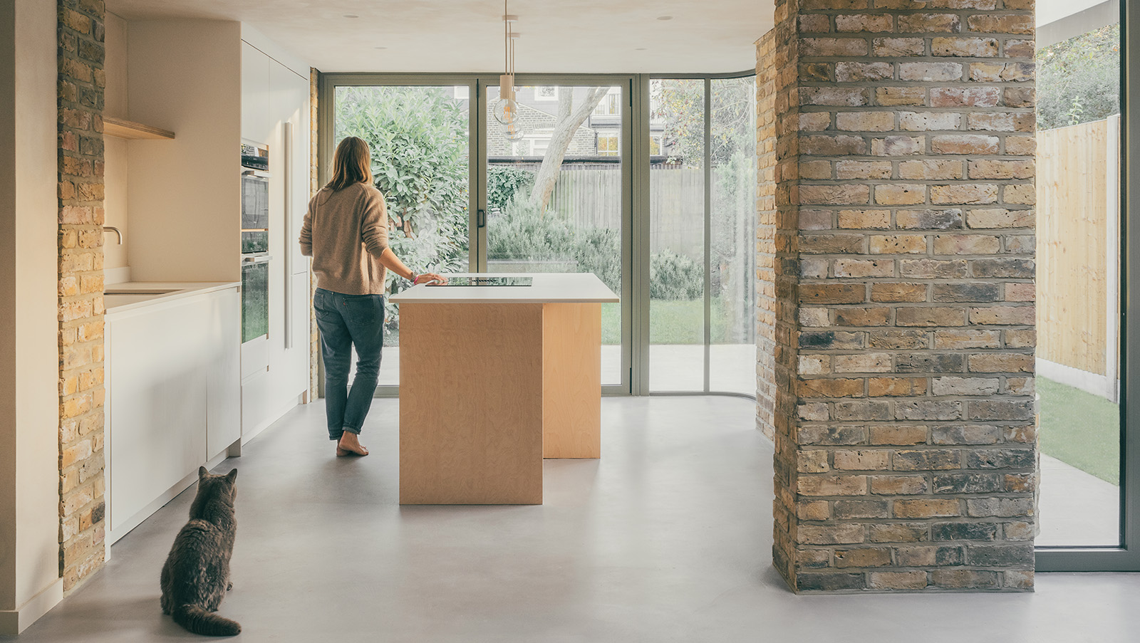 Ashbourne Grove by Pensaer, showing kitchen with woman looking out and cat, the building has natural materials and a minimalist feel - a building moving away from ultra-processed architecture
