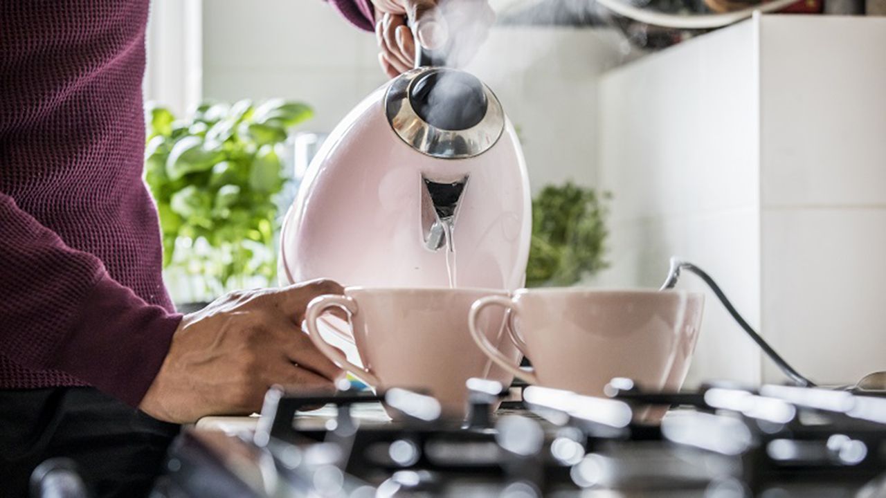 A man wearing a claret colored waffle-textured jumper pouring boiling water out of a pink electric kettle into two pink ceramic coffee cups 