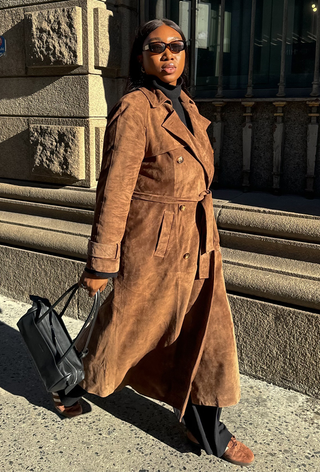 what to wear in 30-degree temperatures is shown in a photo of a woman walking down the sidewalk wearing a brown suede trench coat over a black turtleneck sweater with black trousers, brown suede sneakers, and a black handbag