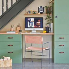 Office area built under the stairs with green cabinets and a pink chair