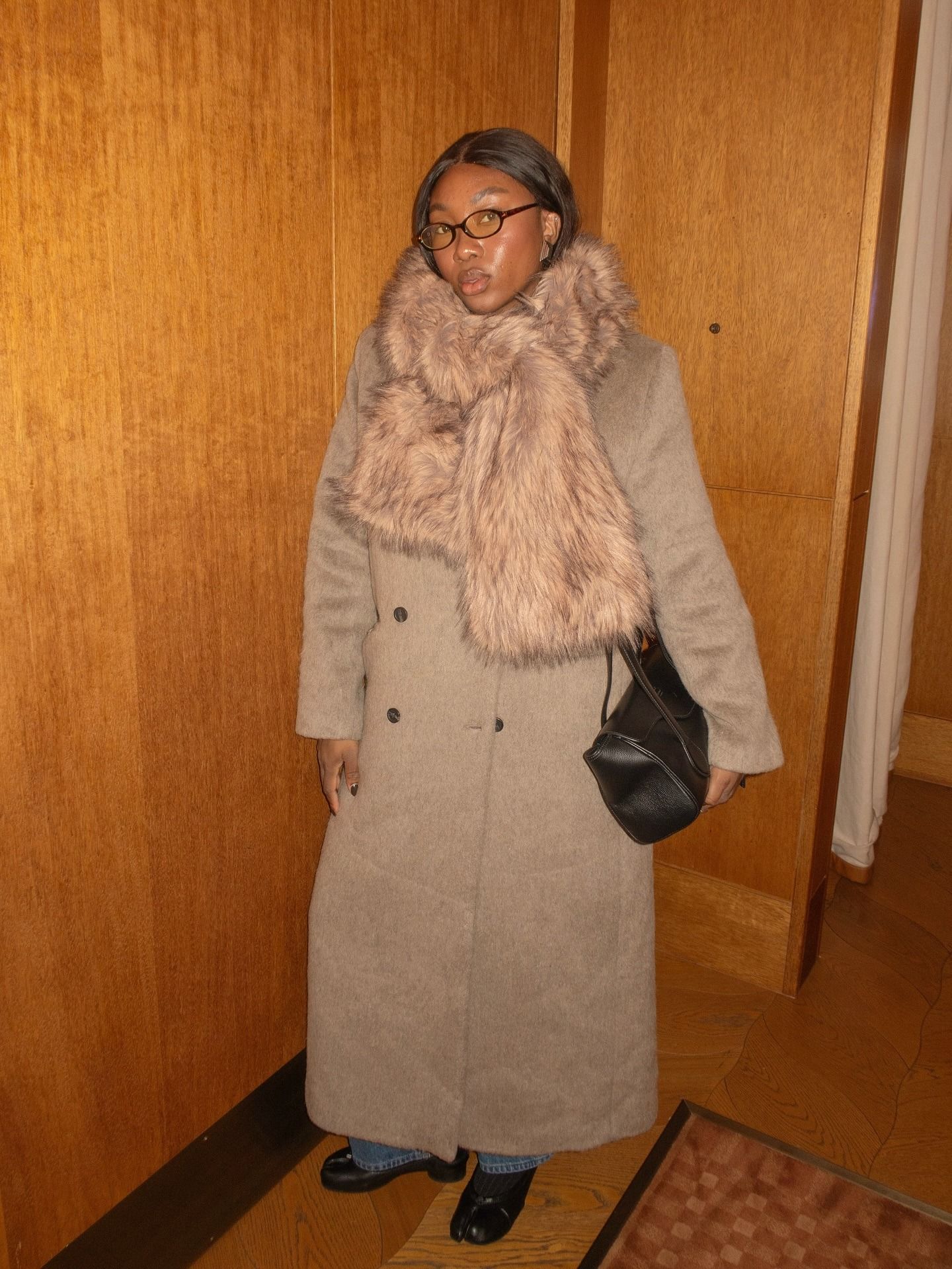 A woman wears a fur scarf, wool coat, black bag, jeans, and loafers.