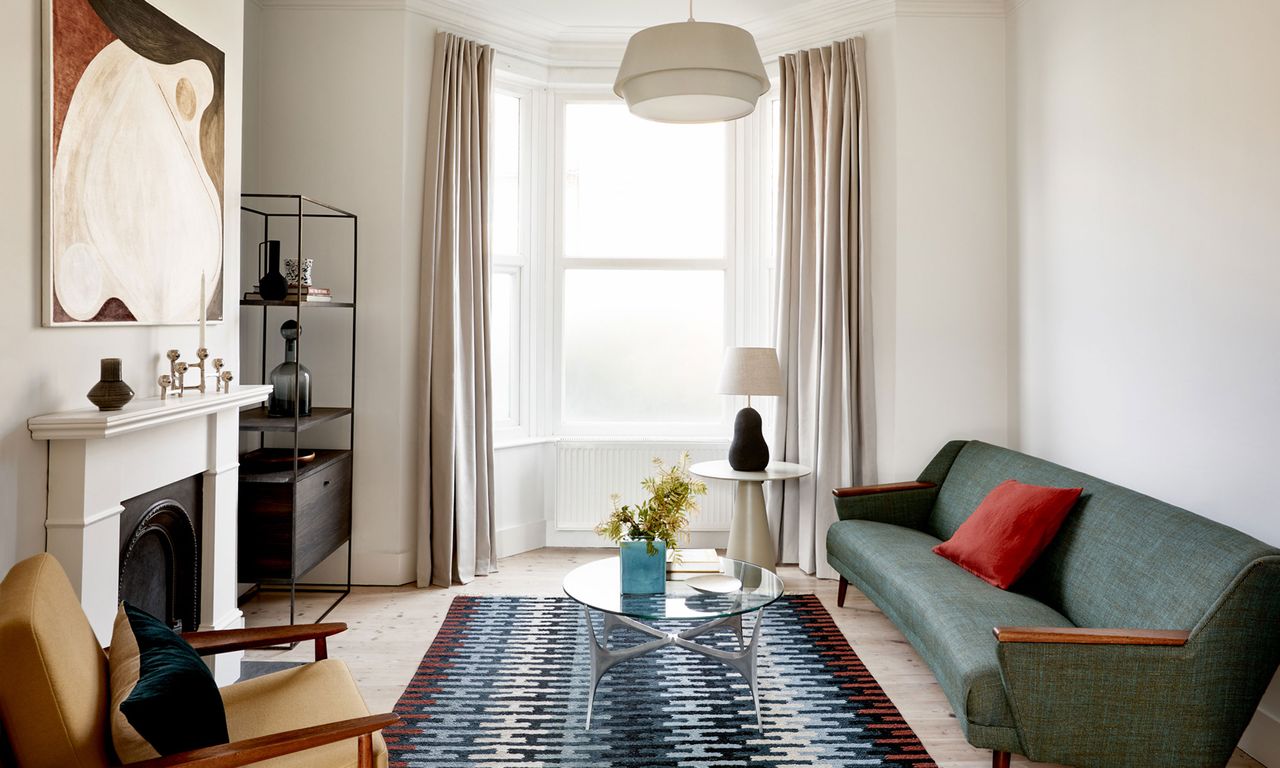 White living room with bay window, full-length curtains, patterned rug, Scandi sofa, glass coffee table, fireplace with candle holder and abstract artwork