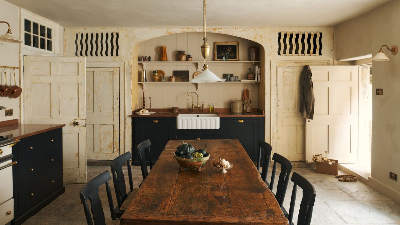 A cream colored kitchen with a large wooden dining table in the middle with chairs on either side as well as a view of the large open shelving above a belfast sink