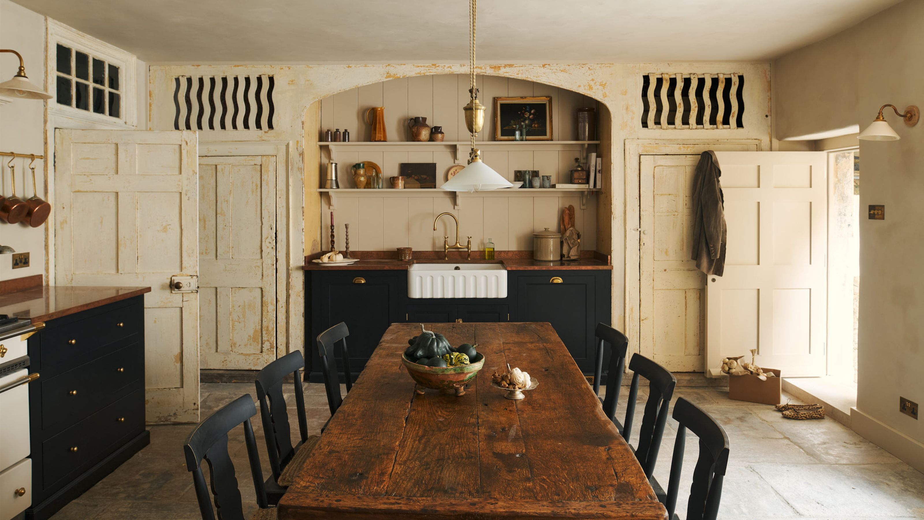 A cream colored kitchen with a large wooden dining table in the middle with chairs on either side as well as a view of the large open shelving above a belfast sink