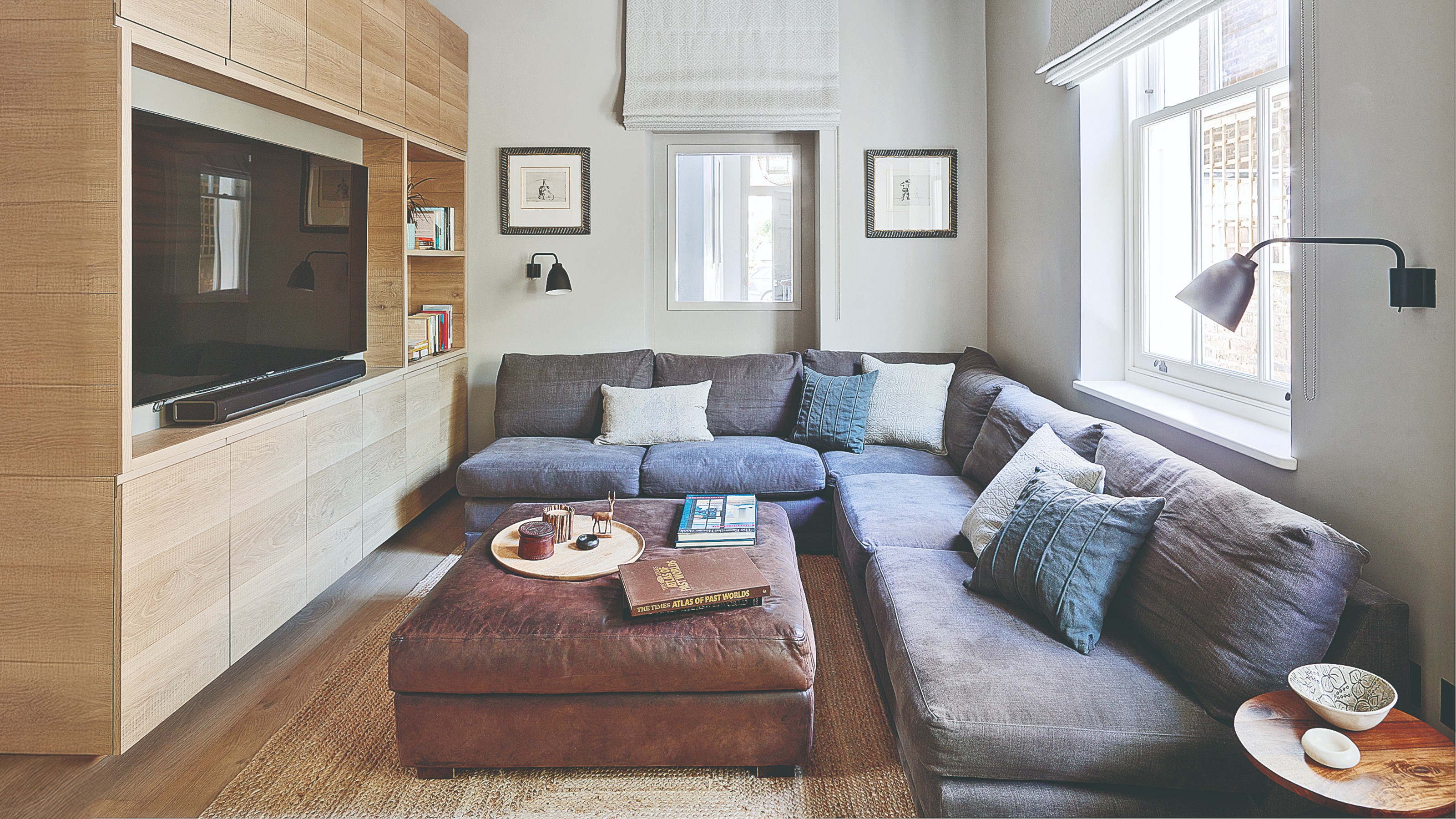 A white TV room with a corner modular sofa in a dark blue linen upholstery with a brown leather ottoman used as a coffee table