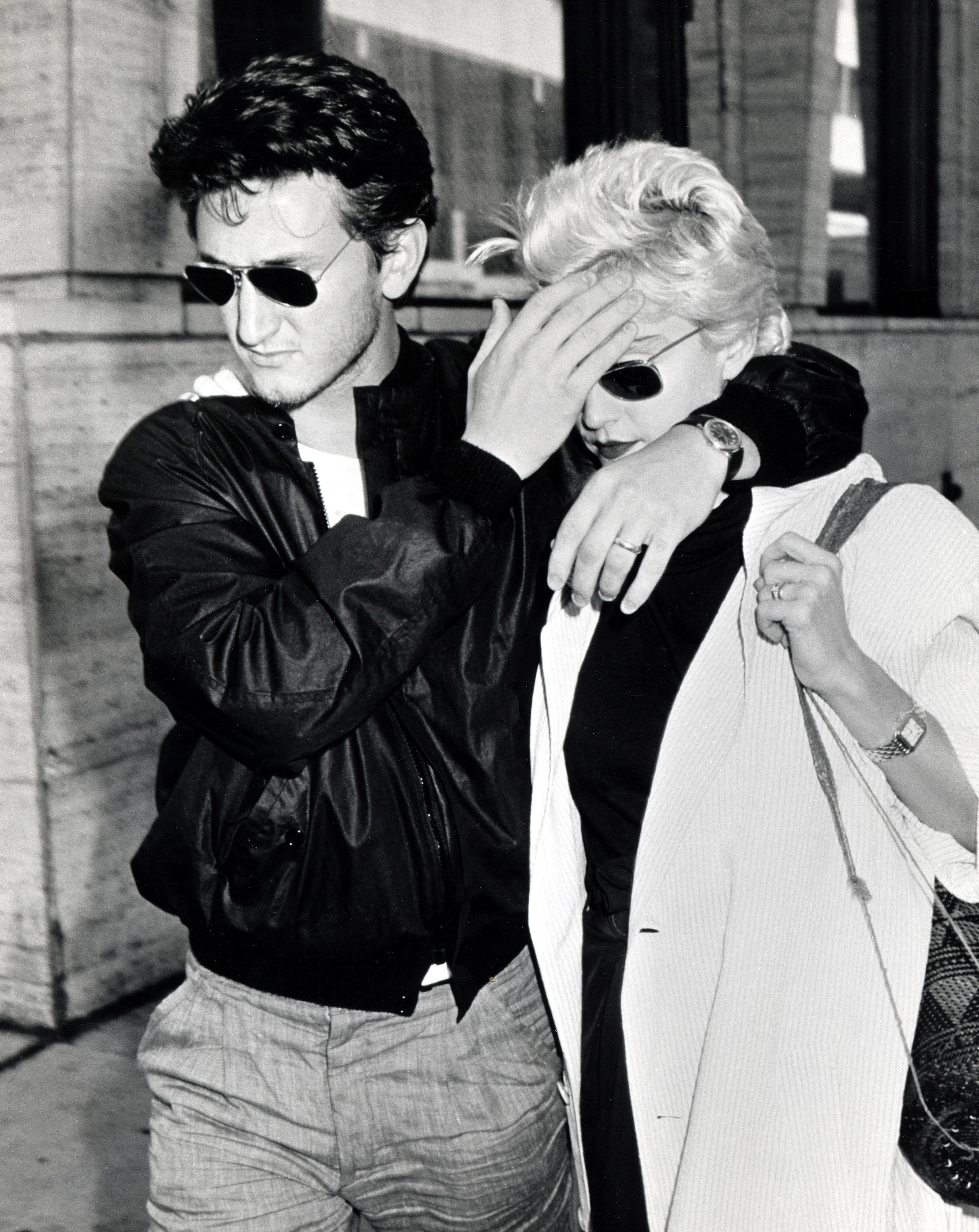 Black and white image of actor Sean Penn wearing a leather jacket and shading the eyes of his female companion from paparazzi flashbulbs