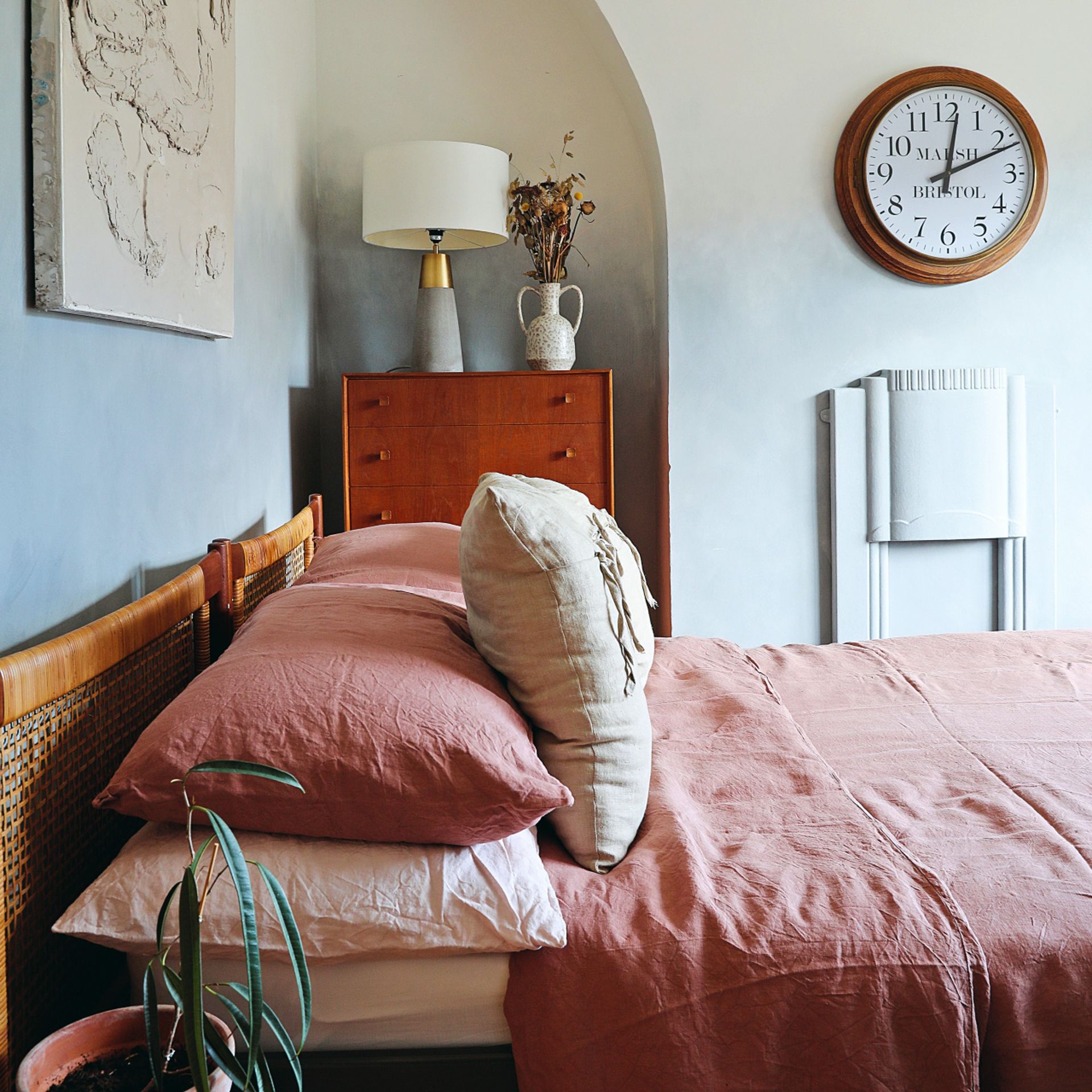 Pink bedding and neutral cushions on double rattan bedframe, hanging wall art, clock, dresser